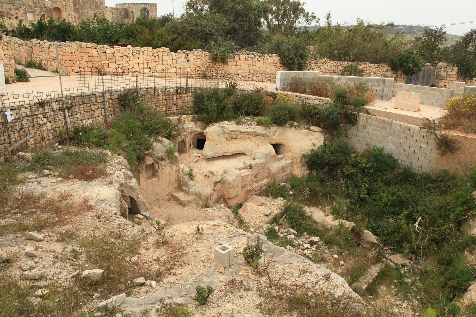 Overview of the Ta' Bistra catacomb site with rock-cut tombs below a railed walkway and rubble walls