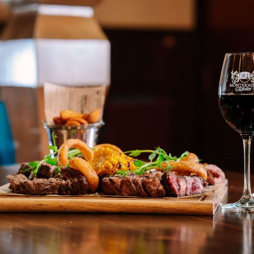 A grilled steak platter with onion rings and a glass of red wine on a wooden table