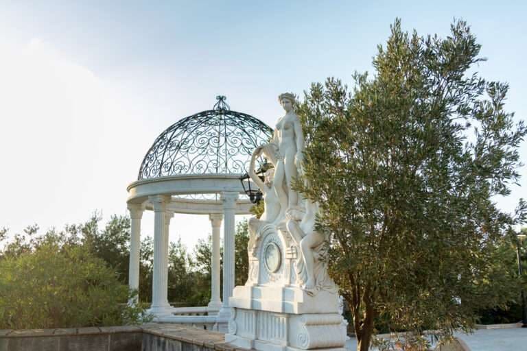A white domed garden pavilion with a statue, beside an olive tree