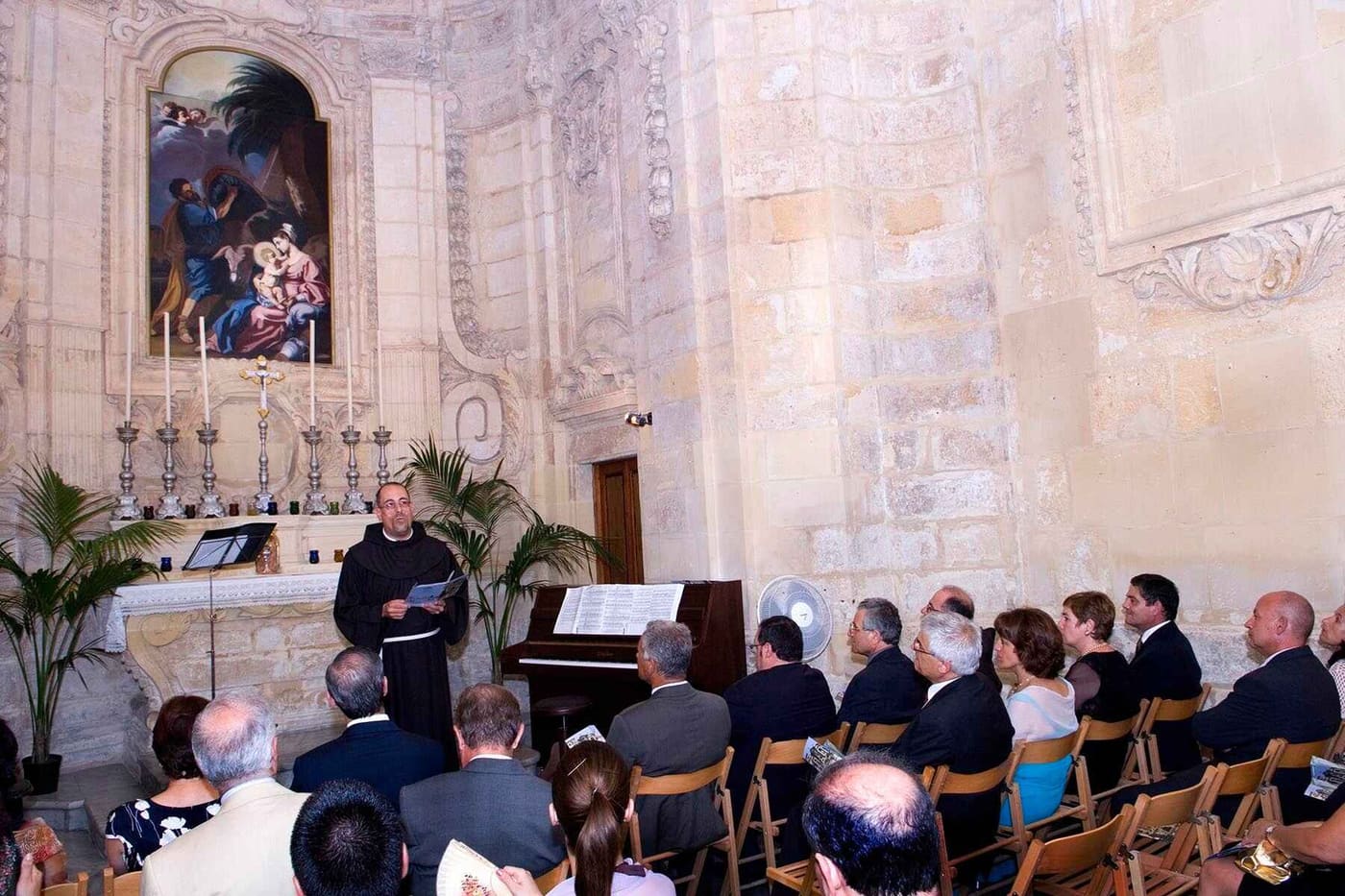 A small congregation seated in the baroque chapel with the altar painting behind a priest