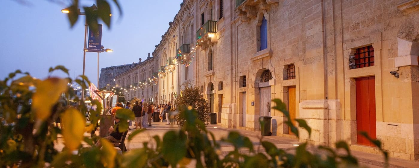 Evening view along the lit stone frontage of the Valletta Waterfront with people strolling