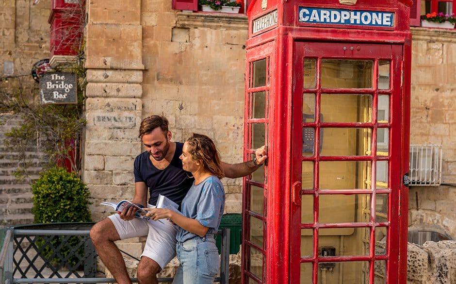A couple looking at a guidebook beside a red telephone box on a Valletta street