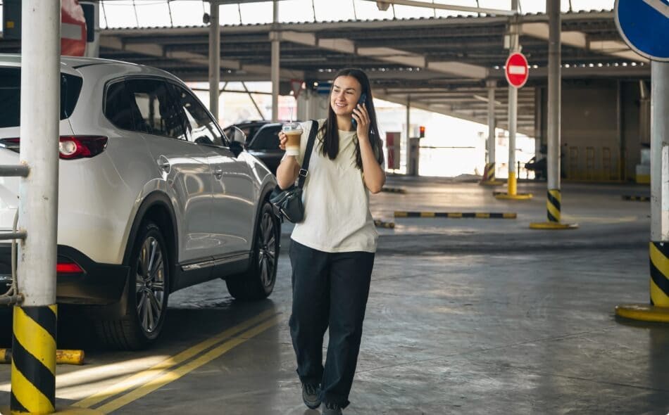 A woman on the phone walking through a covered car park beside a white car