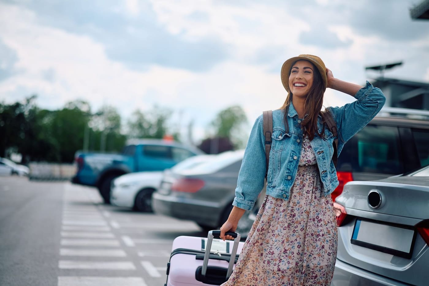 A smiling traveller in a straw hat pulling a pink suitcase through a car park