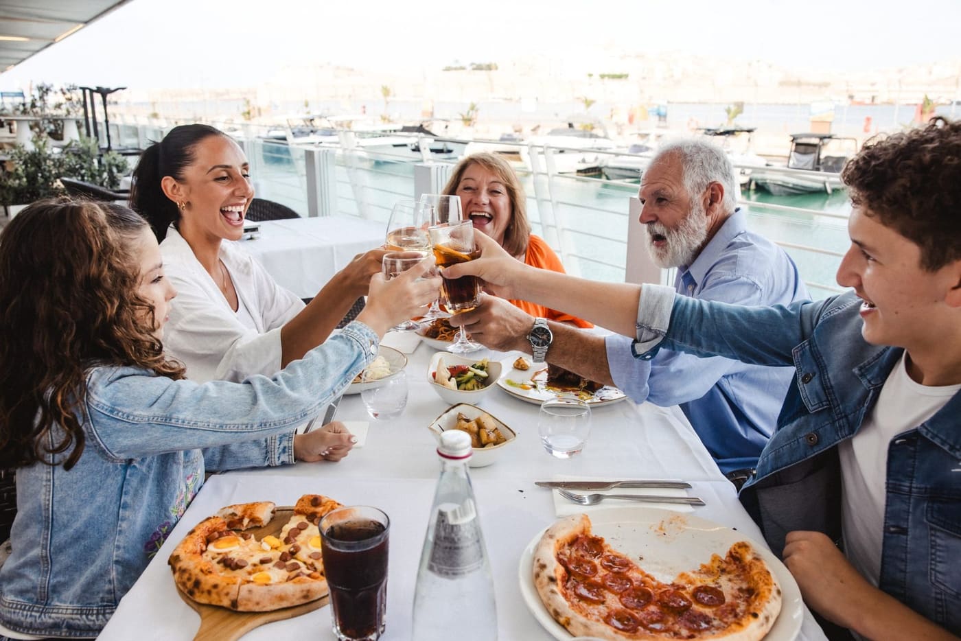A family toasting around a table with pizzas on a harbourside terrace